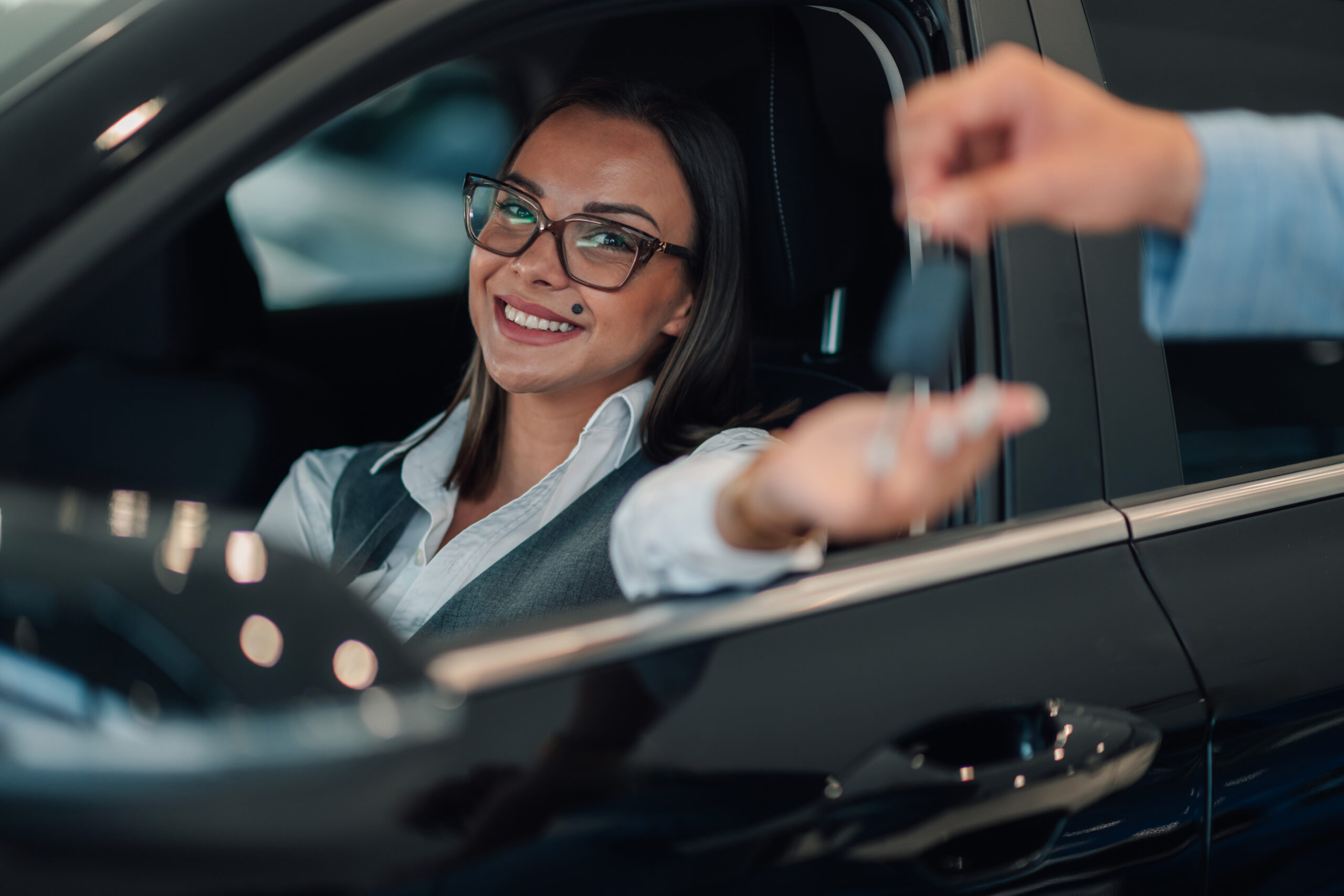 A woman, smiling broadly, leans forward from inside her car to reach for keys being handed over by another person, capturing a moment of joy and accomplishment in car acquisition.
Adonis Rent a Car